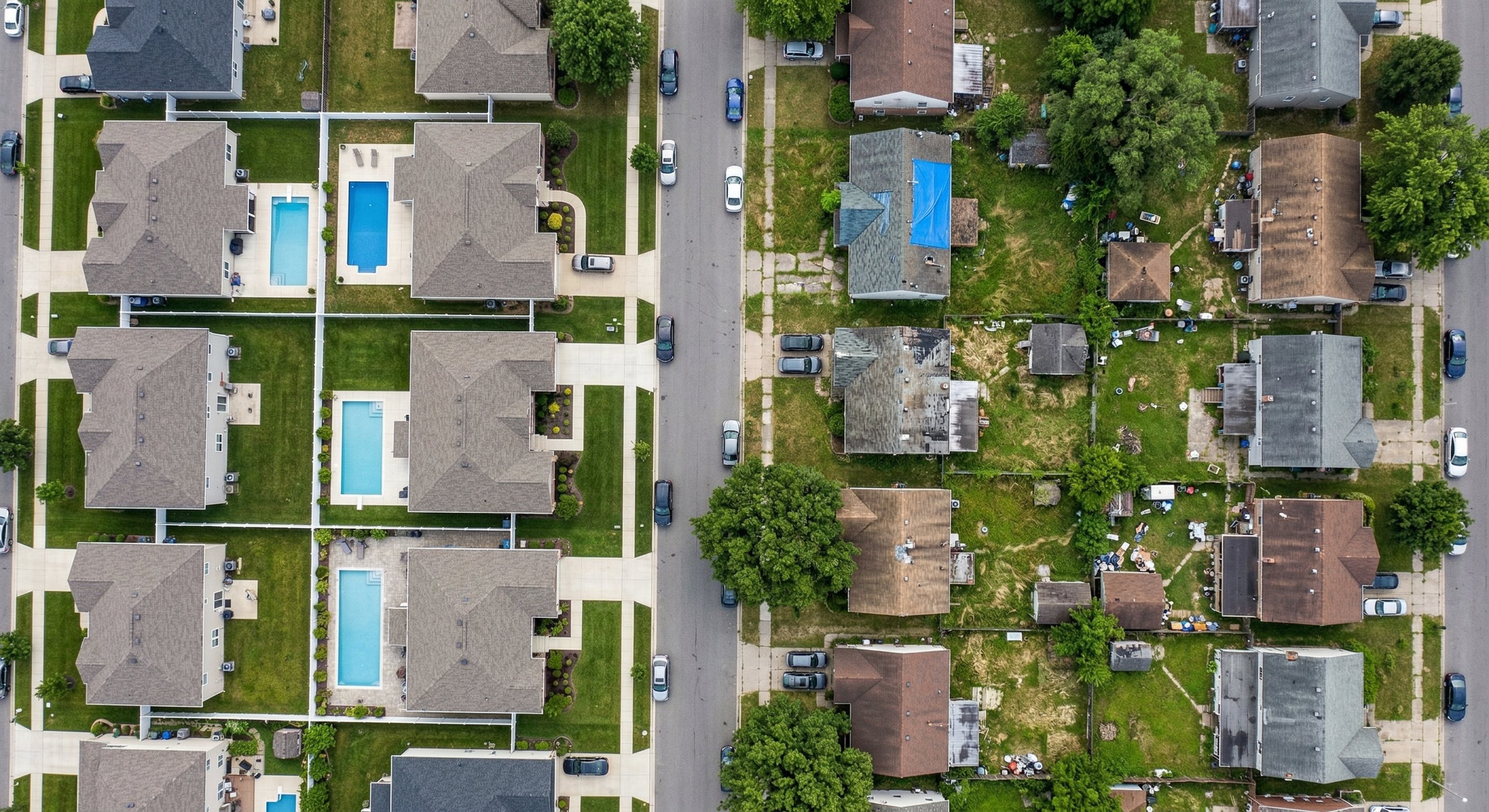 An aerial drone photograph from 400 feet, looking straight down at a mid-sized American city's residential neighborhood with varied housing density — a mix of well-maintained homes and visibly neglected properties. Some properties have overgrown yards or aging roofs visible from above. Midday sun, real estate aerial photography, DJI Mavic 3.